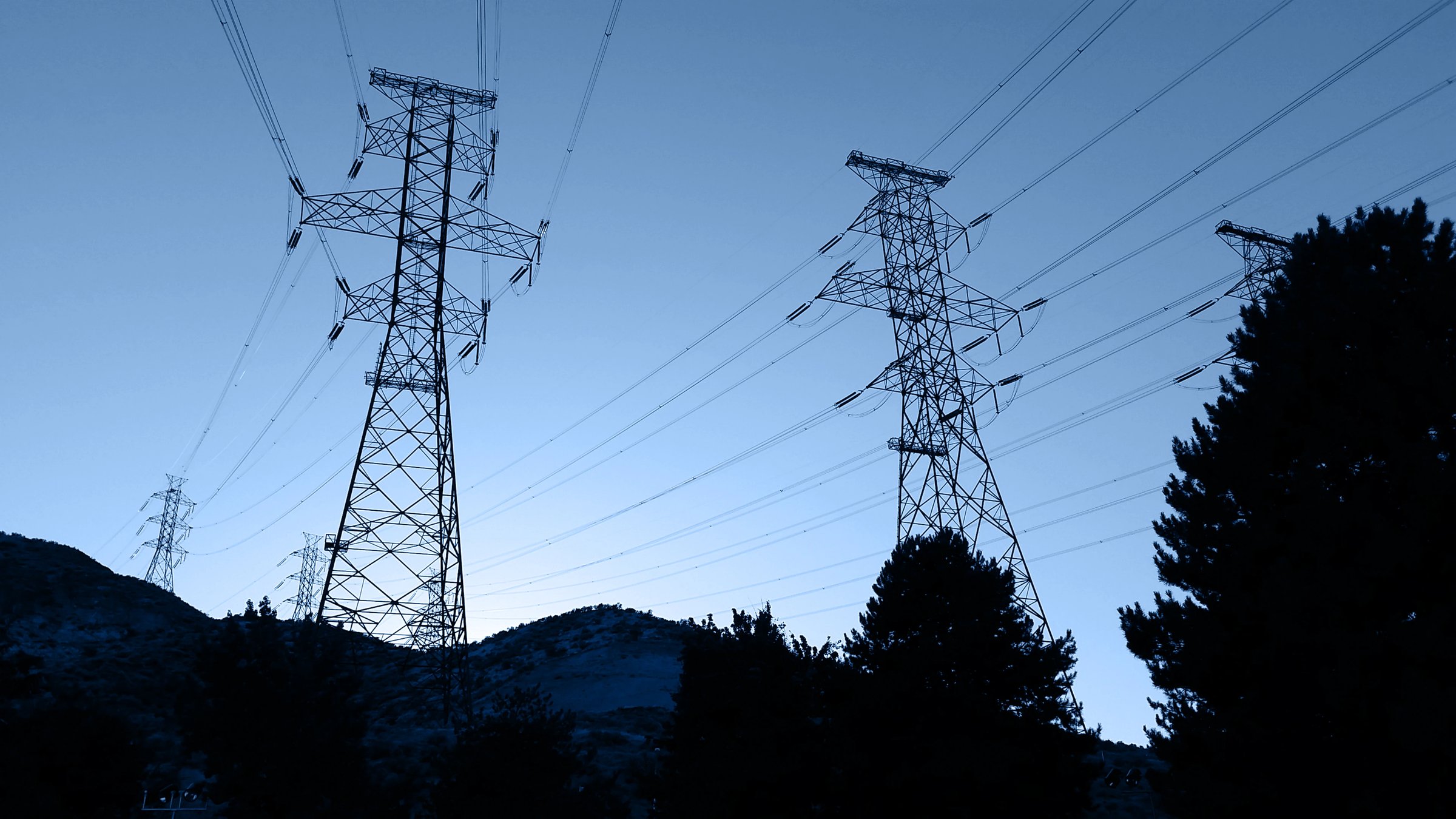 Steel transmission towers and high-voltage lines silhouetted against a twilight sky above hills and trees.